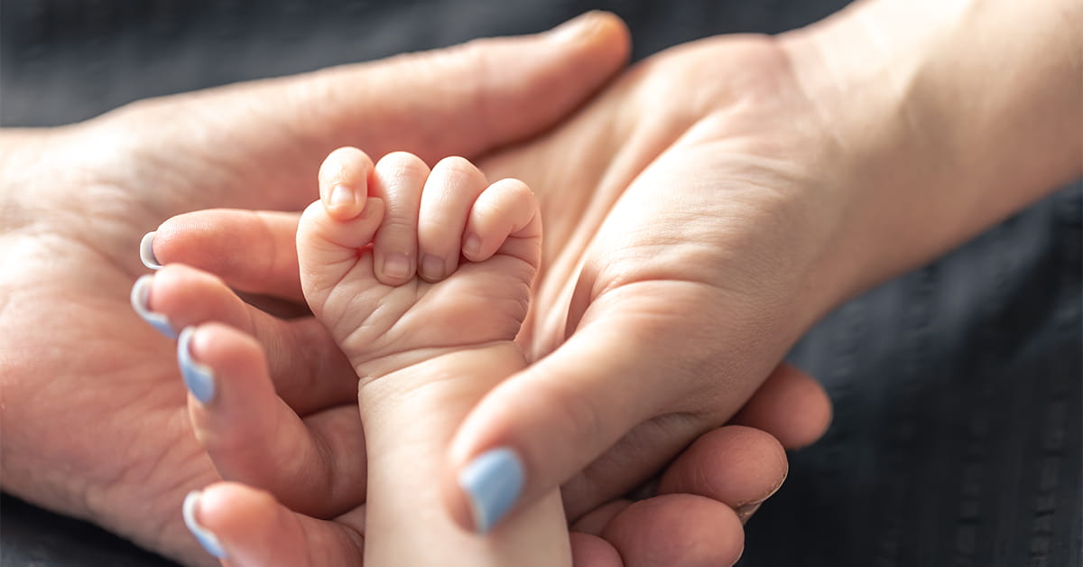 Adult hand with blue nail polish gently cradles a baby's tiny fist, showing a tender bond.