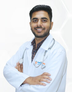 Smiling male doctor in a white coat with a stethoscope, arms crossed, standing against a light geometric background.