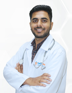 Smiling male doctor in a white coat with a stethoscope, arms crossed, standing against a light geometric background.